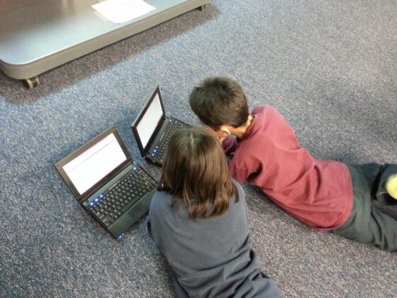 Due bambini di spalle, su una moquette grigia, mentre usano i loro laptop