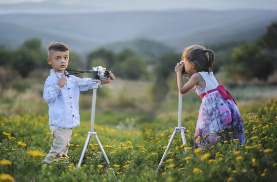 Un bambino e una bambina in mezzo a un prato, ognuno davanti ad un cavalletto con una macchina fotografica 