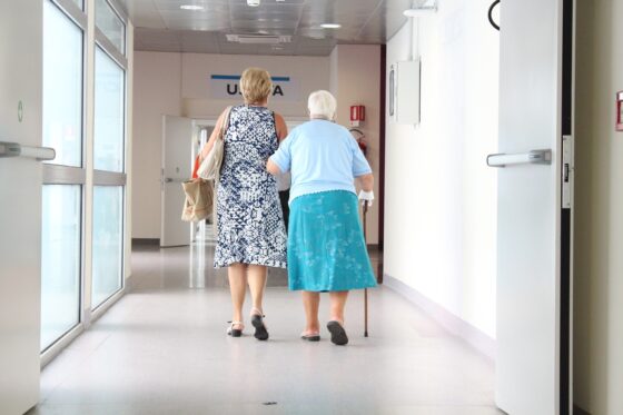Un'anziana di spalle in un corridoio d'ospedale cammina appoggiandosi ad una donna più giovane