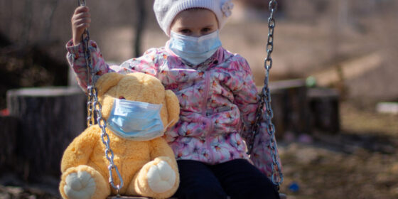 Una bambina su un'altalena con il suo orsetto di peluche, entrambi con la mascherina